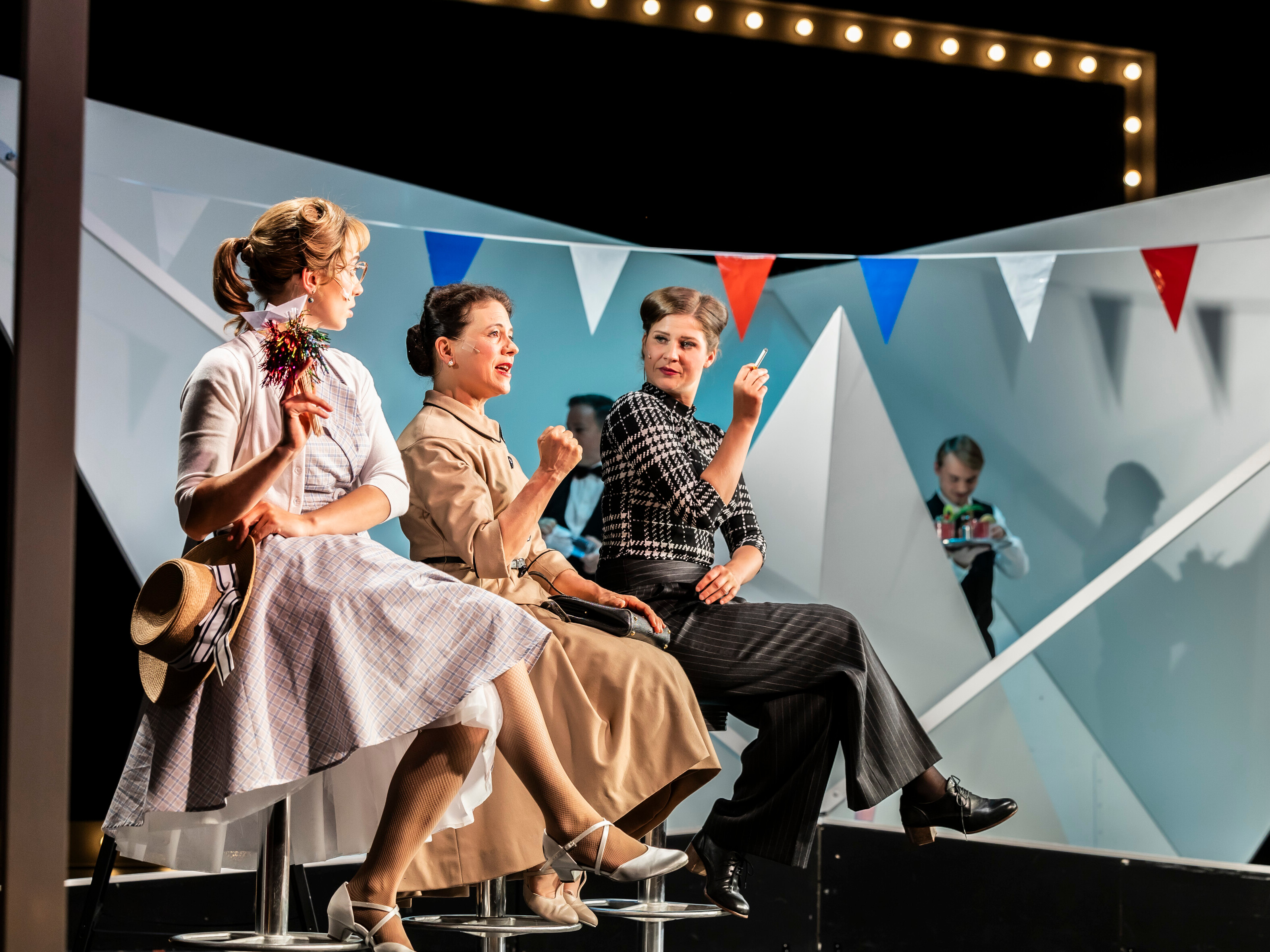 Drei Frauen in Kostümen der 60er Jahre sitzen auf Hockern und unterhalten sich, während eine Figur im Hintergrund Getränke vorbereitet. Szene aus der Aufführung "Petticoat und Minirock" im Theater Lüneburg, Foto: Jochen Quast.